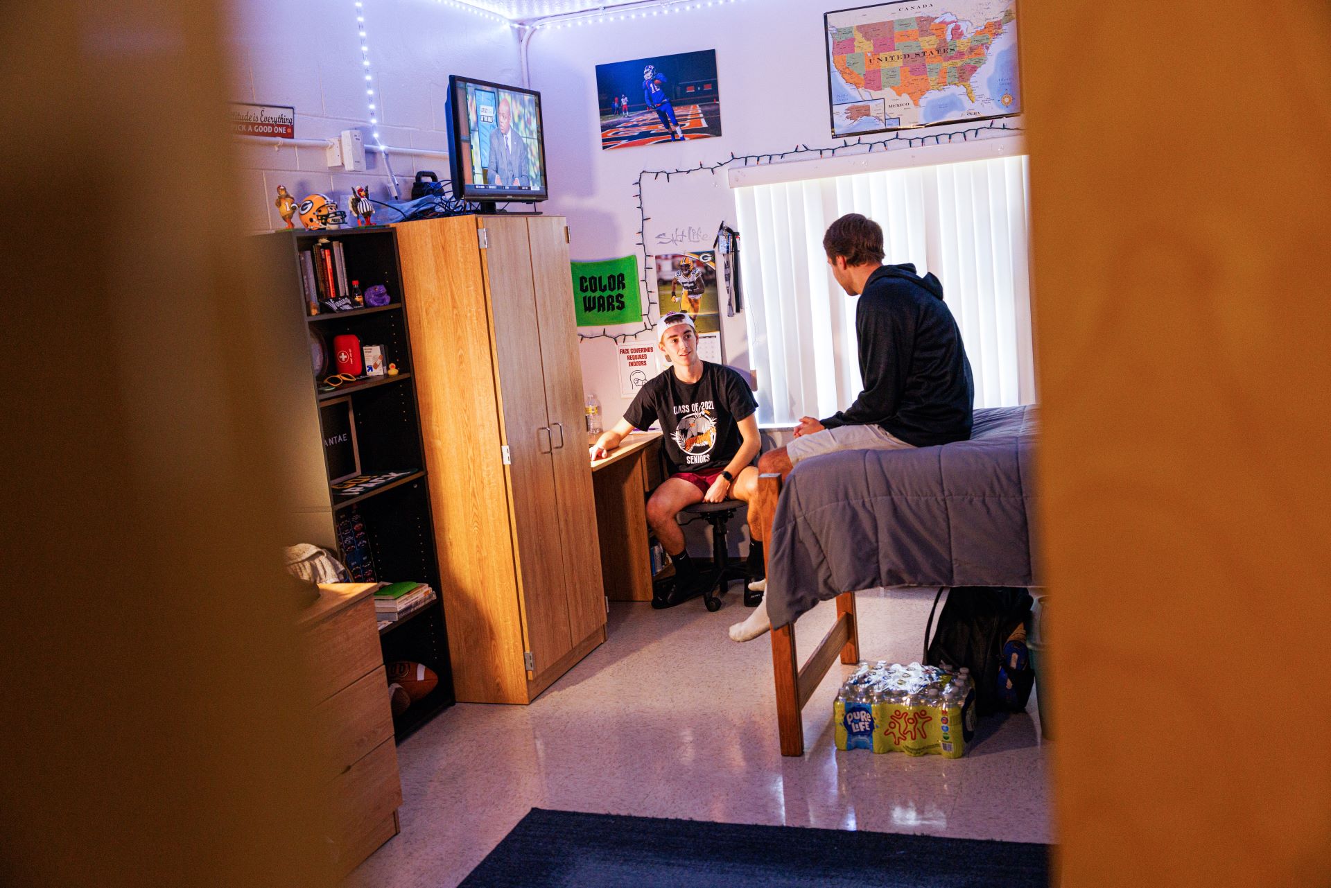 Two college students chatting in a dorm room, viewed through an open door; one sitting at a desk and the other on a bed, with a TV showing a news program in the background
